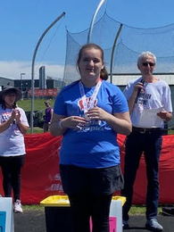A female student smiles while standing on an outdoor awards podium, wearing a blue Special Olympics t-shirt and holding a medal around her neck. An announcer and a volunteer applaud in the background under a clear blue sky.